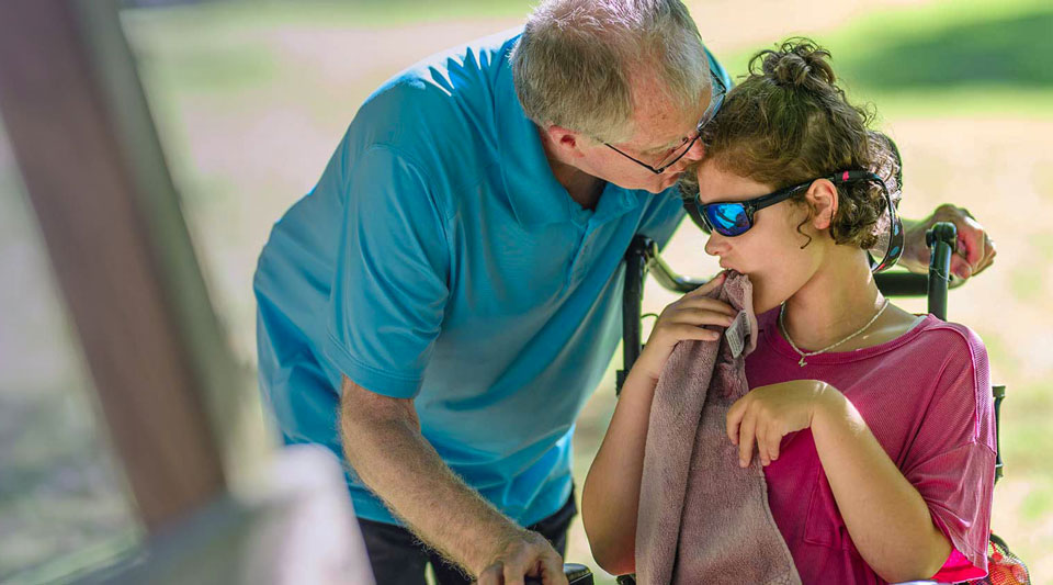 Older man in blue shirt leans toward young person in wheelchair with sunglasses.