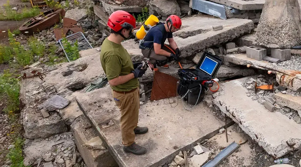 Two people in red helmets operate equipment amongst concrete debris.